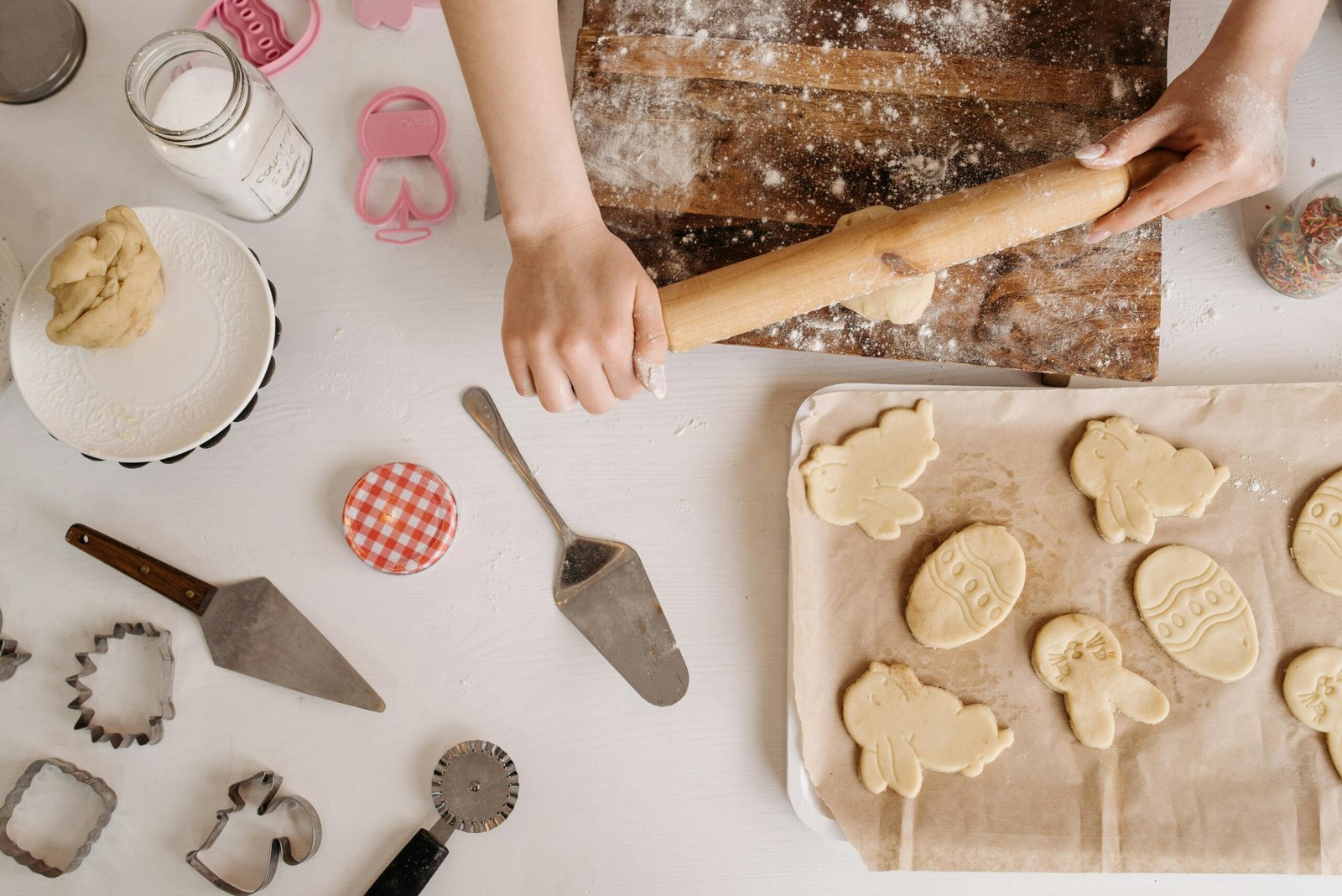 Hands rolling dough for cookies with baking tools and ingredients on a wooden table.